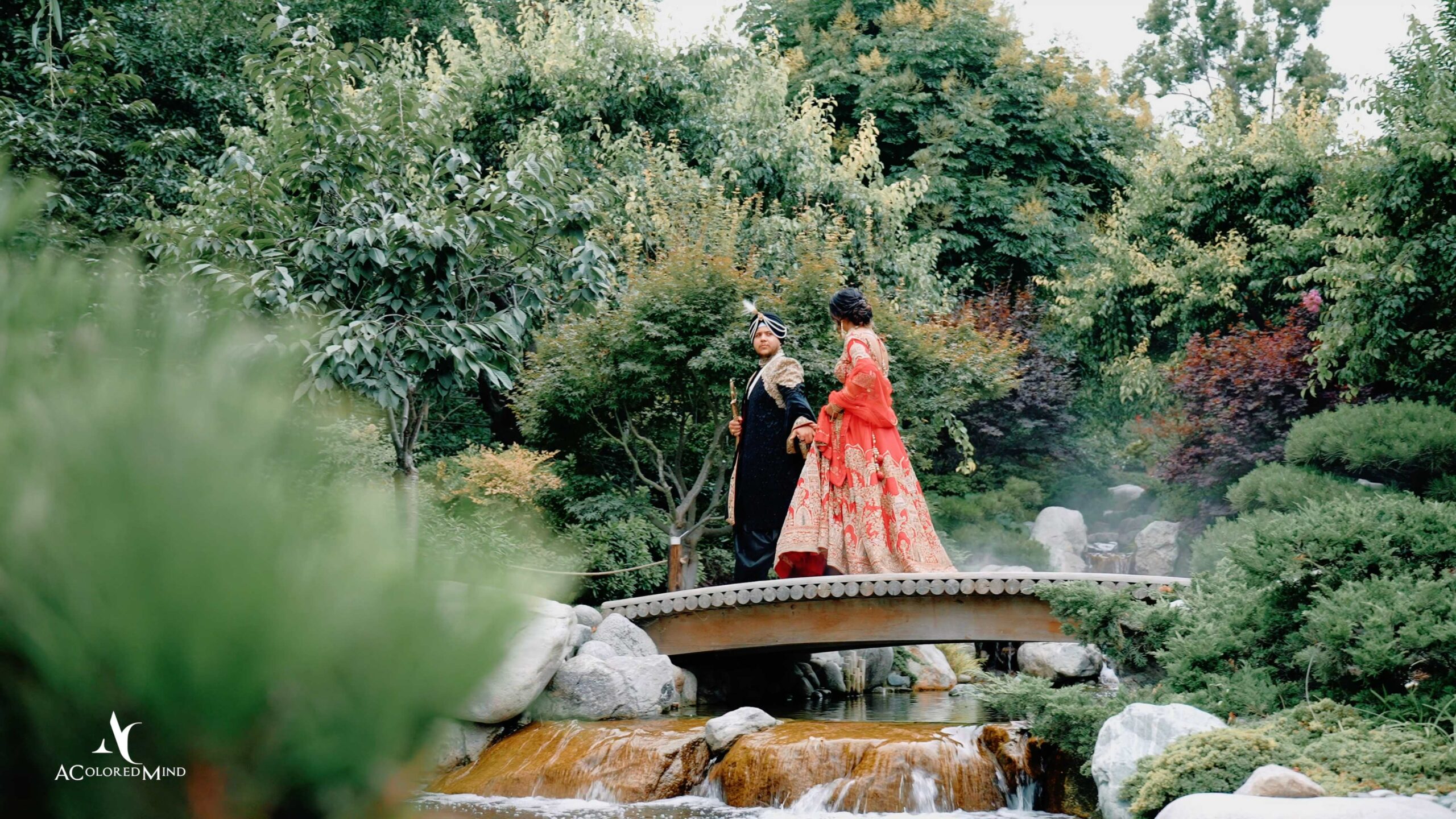 Bride and groom in traditional Indian Sikh attire holding hands on a garden bridge surrounded by lush greenery and trees.