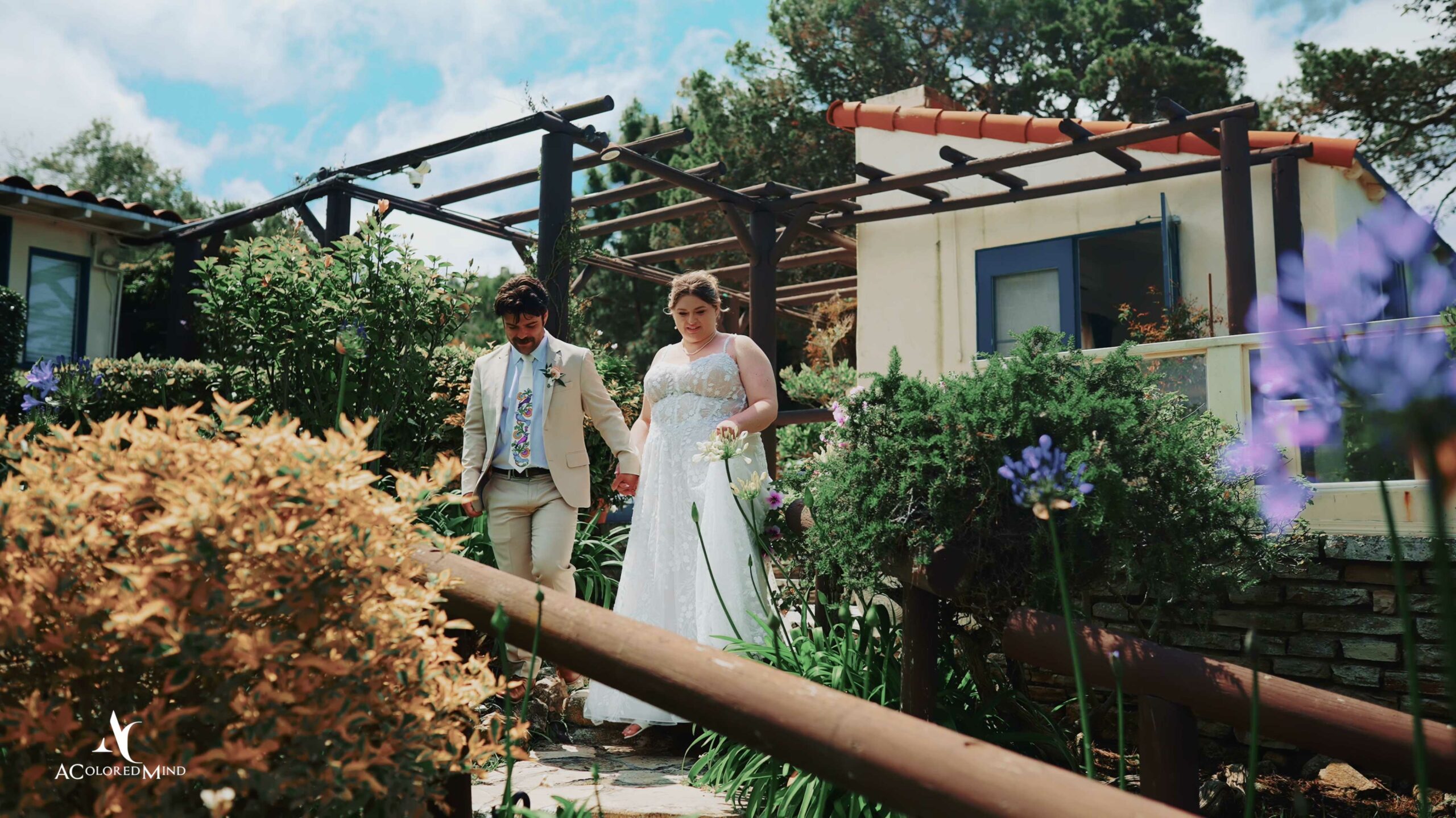 Bride and groom walk hand-in-hand through lush garden path at La Venta Inn, surrounded by blooming flowers and greenery.