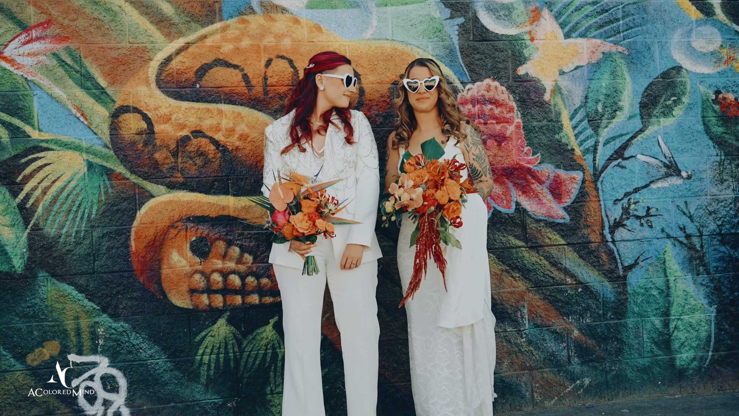 Two brides wearing heart-shaped sunglasses in front of a colorful mural at The Millwick in the Arts District, Los Angeles—perfect for modern LGBTQ+ wedding videography.