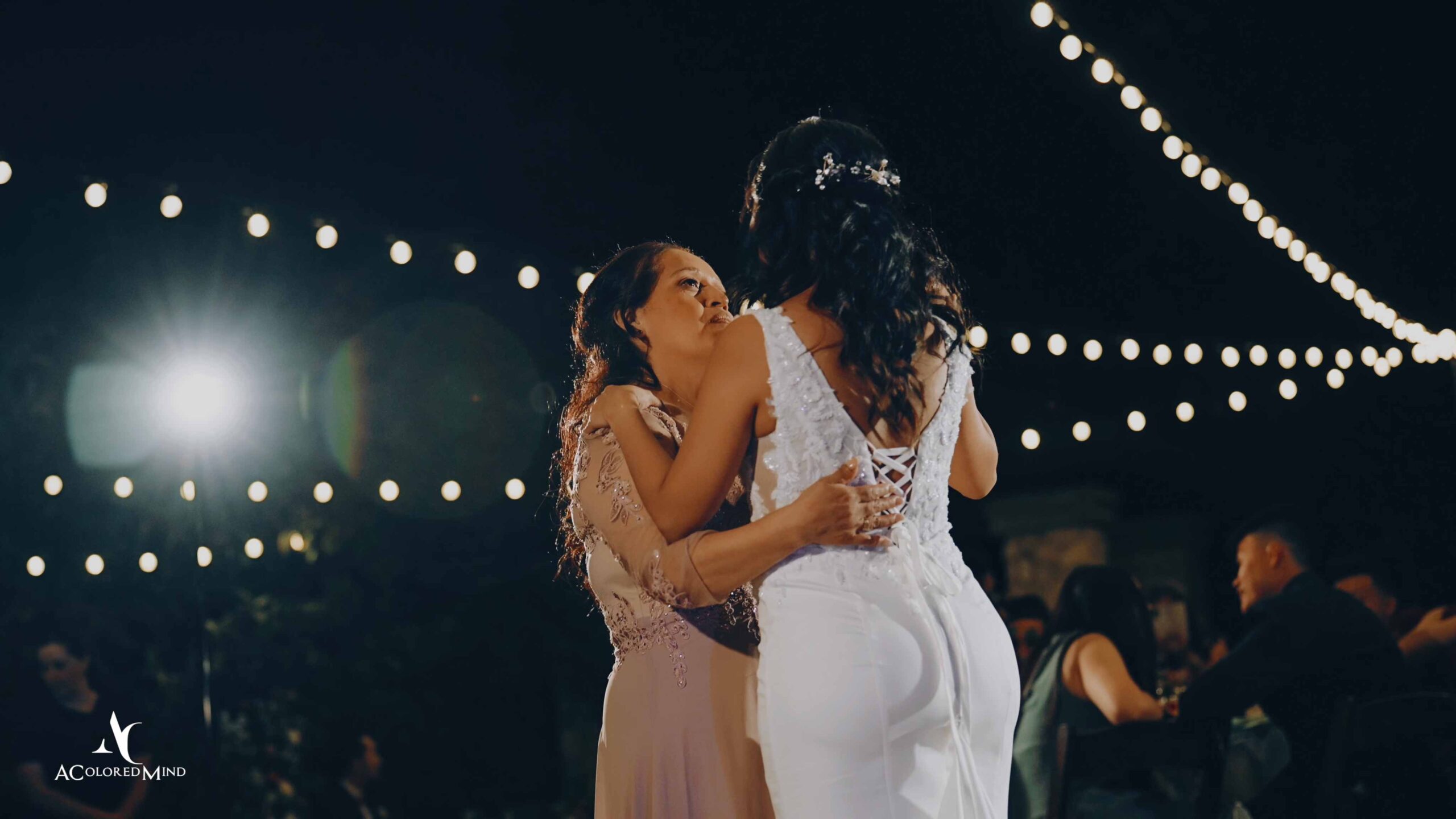 A bride in a white lace wedding dress shares an emotional dance with her mother during an outdoor Los Angeles wedding reception illuminated by overhead string lights.