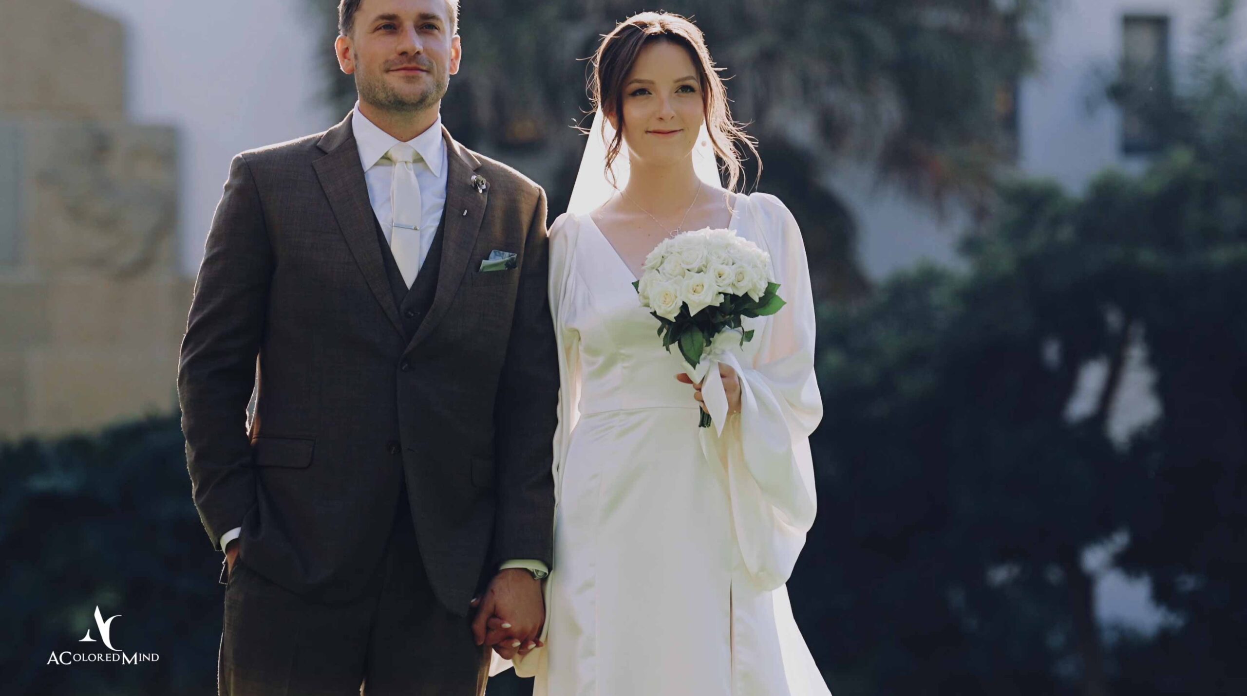 Bride in a flowing long-sleeve white dress holding a white rose bouquet, standing outdoors holding hands with her groom in a brown suit.