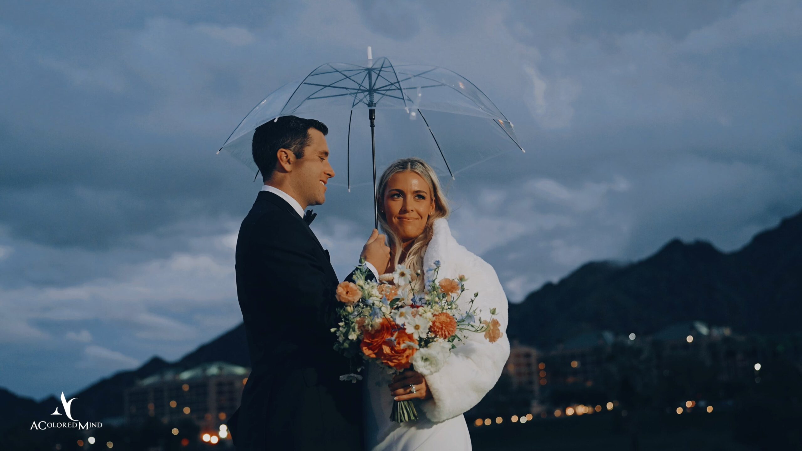 Bride and groom standing under a clear umbrella at twilight with mountains in the background during their Indian Wells Golf Resort wedding.