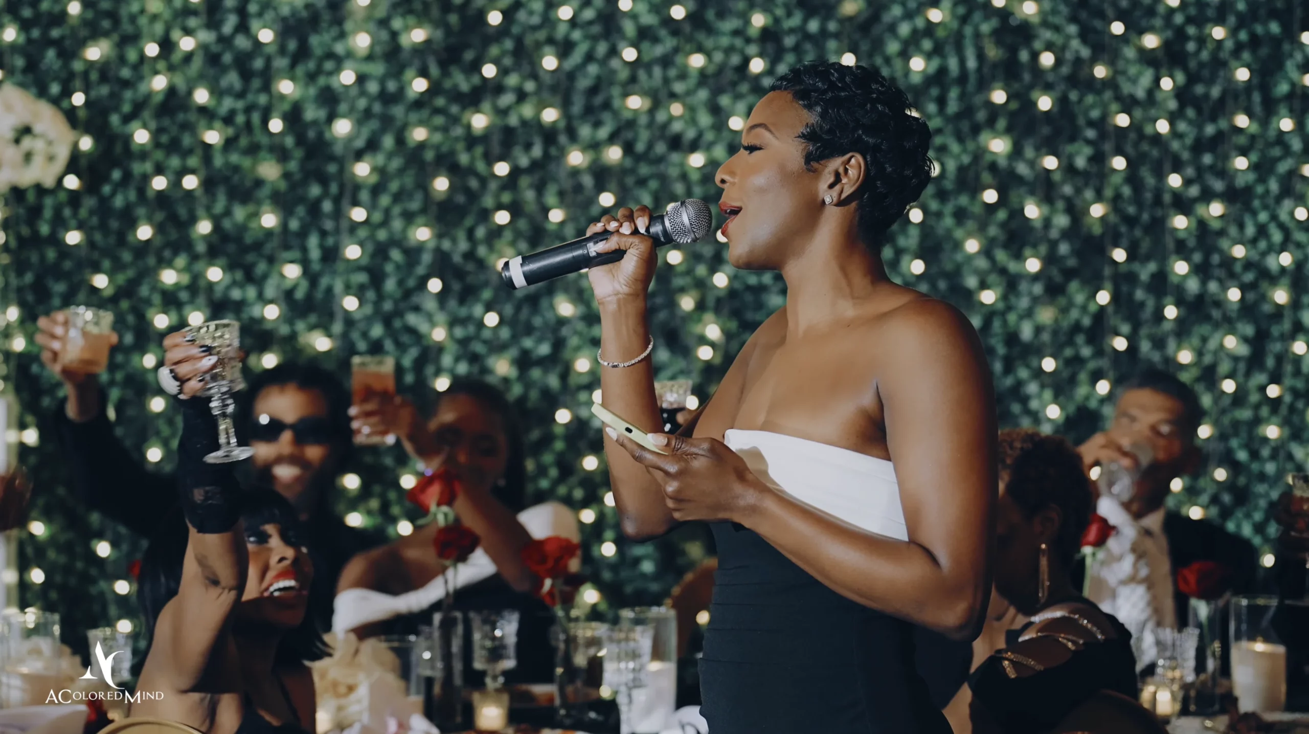 A wedding guest in a strapless black and white dress gives a heartfelt reception speech with a microphone at The Oviatt in Downtown Los Angeles, while seated guests raise their glasses to toast in front of a green boxwood wall illuminated by fairy lights
